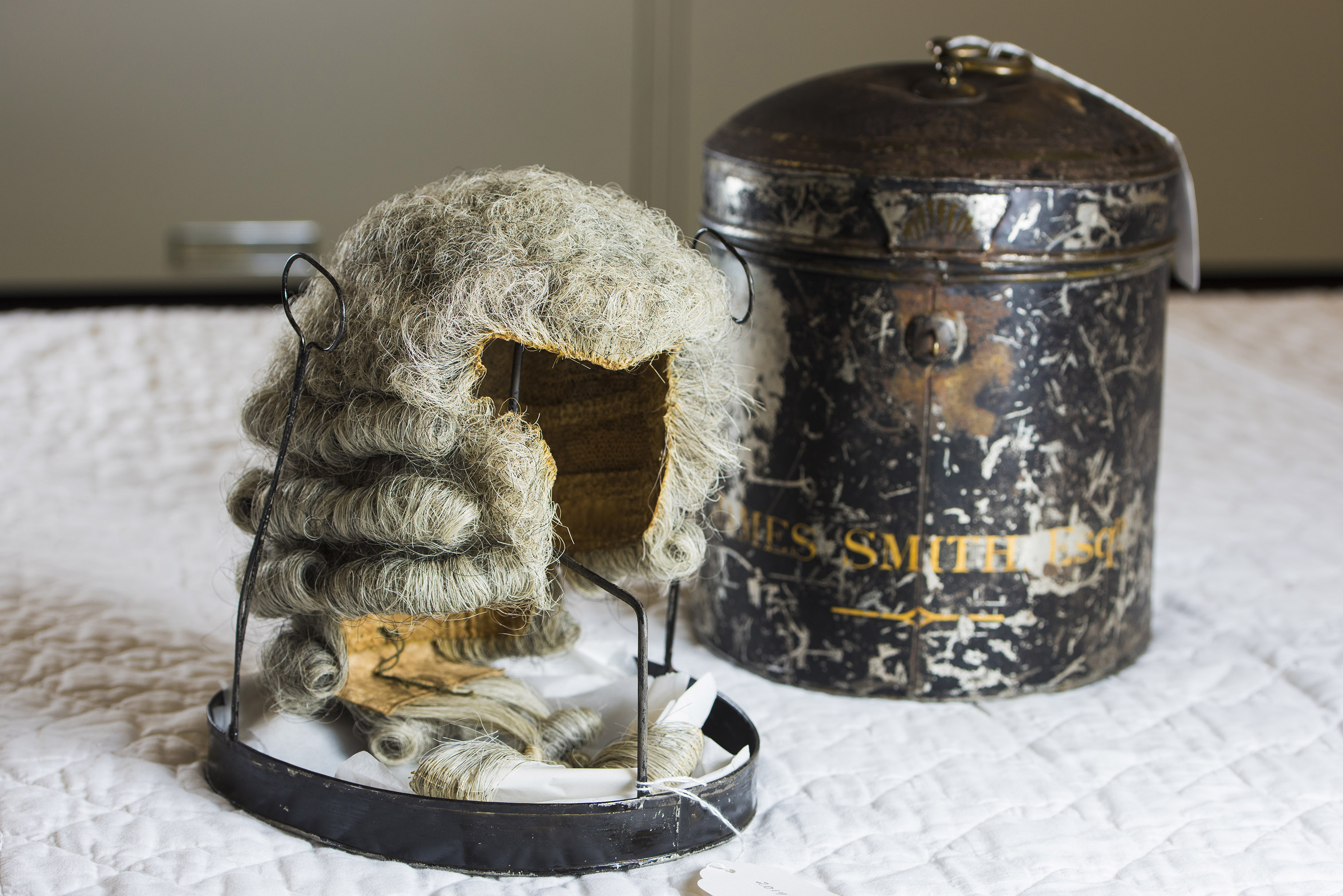 A grey, powdered wig on a wooden head form, displayed next to a fading, black metal canister.