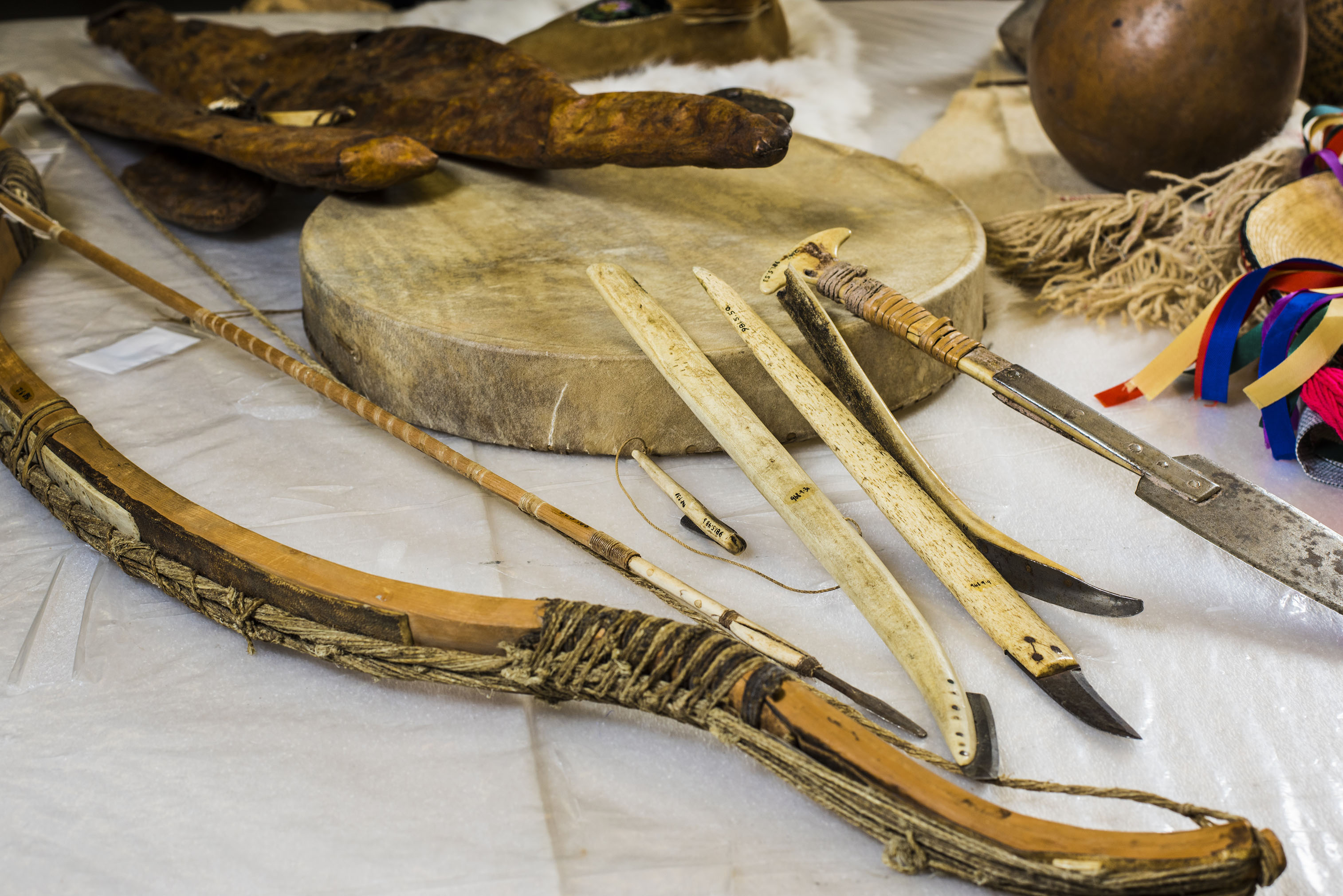Artifacts on a covered table, including a bow, a round hand drum, and a sharp metal tool with a covered handle.