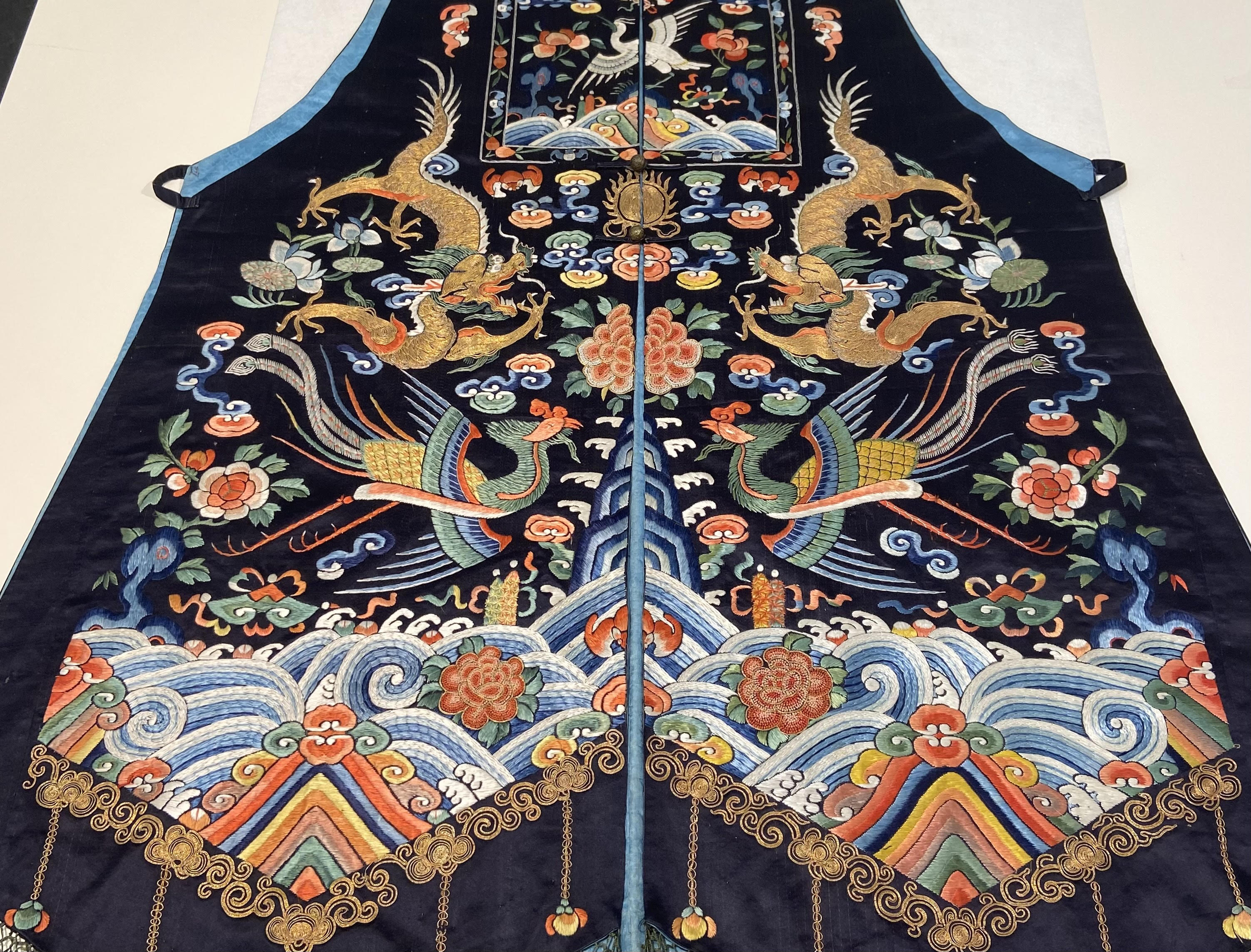 A highly detailed embroidered vest rests flat on a white surface. The sides of the robe are mirrored, and feature, dragons, birds, flowers, and waves, all presented in a multitude of colours.