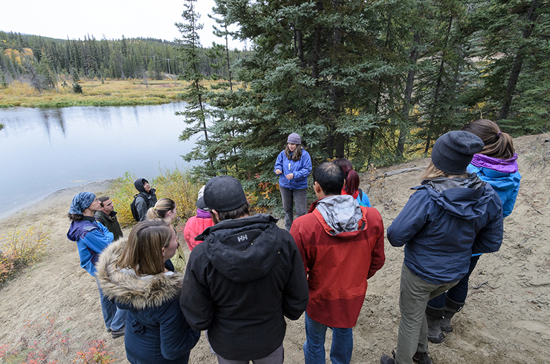 A group of seven or eight students majoring in northern systems listen to instruction on a riverbank in the Yukon.