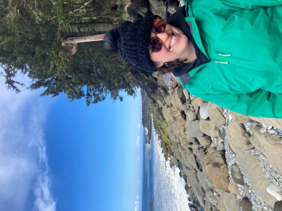 A photo of Allie in a green sweater and toque in front of a rocky shore.