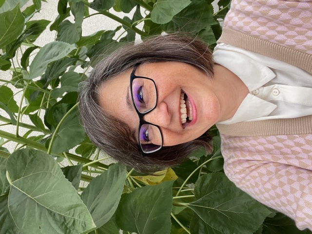 A photo of Cristine in a pink sweater vest in front of leaves.
