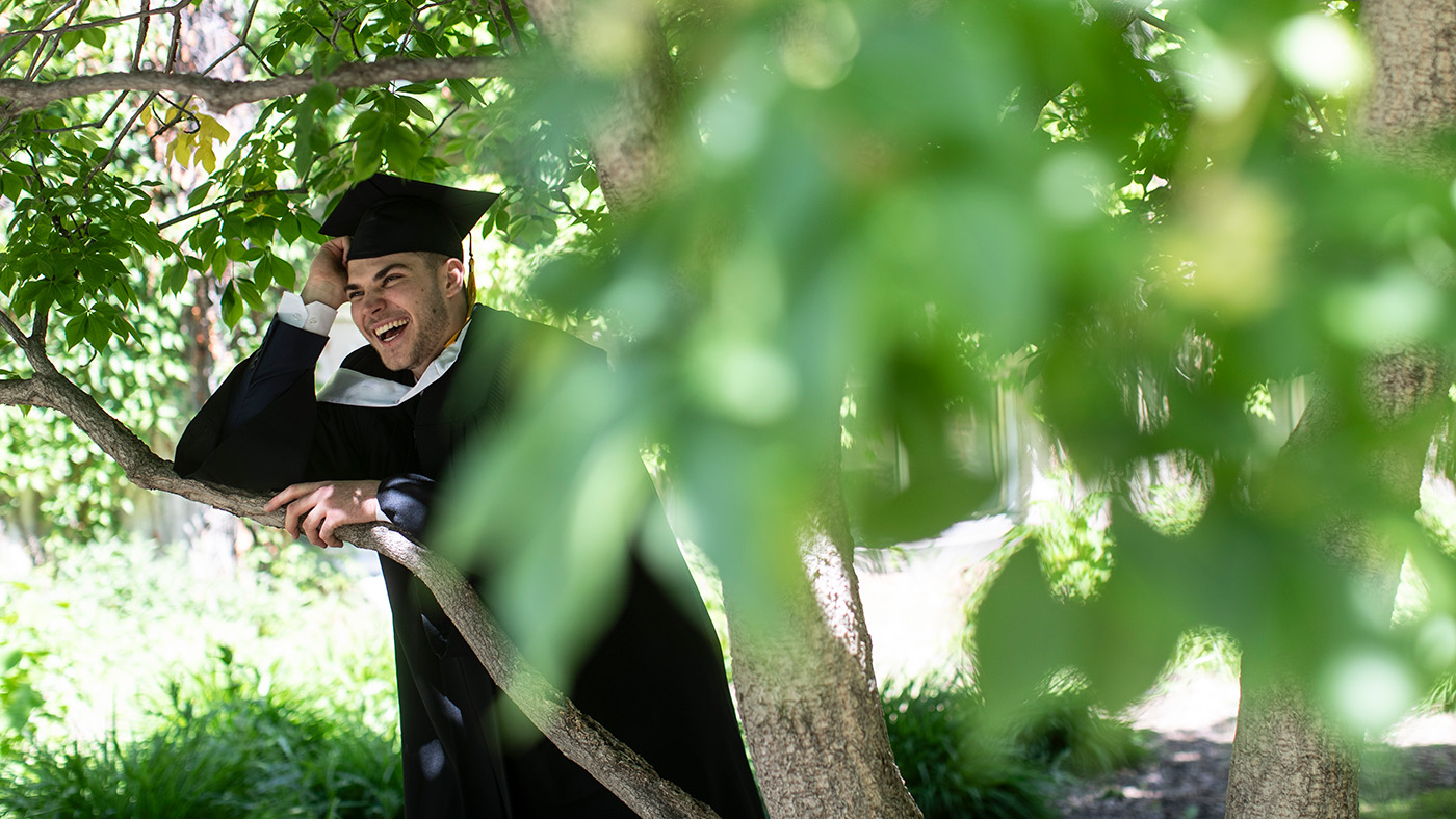 Graduand smiling while posing for photos in green low tree.