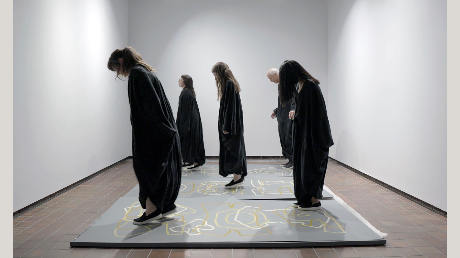 A group of 5 people wearing dark cloaks and matching shoes are facing left in a specific formation. They look like they might be dancing. They are on a constructed wood floor that is painted with abstract patterns and shapes.