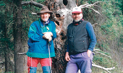 Garry Gibson and Doc Larson leaning against a tree in a wooded area while wearing outdoors clothing.