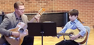 A student and teacher sitting and playing their guitars while looking at music.