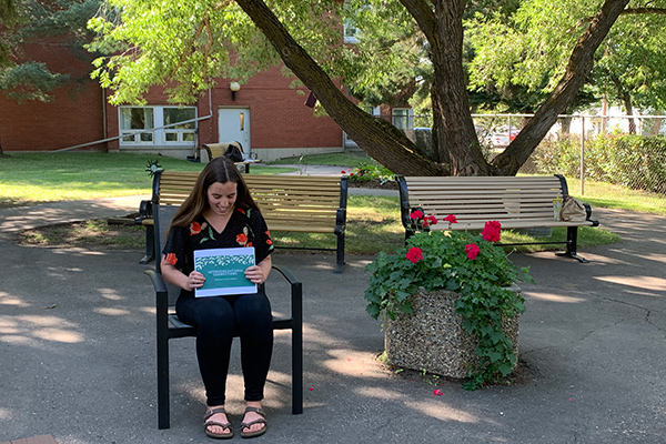 Jaden de Waal sits at a bench in the courtyard at Memory Lane holding up a paper presentation of her summer project.