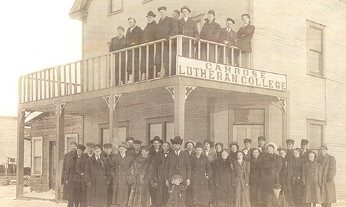 Sepia photo of people standing in front of Heather Brae Hotel and on the balcony of the hotel. The sign "Camrose Lutheran College" hangs on the front.