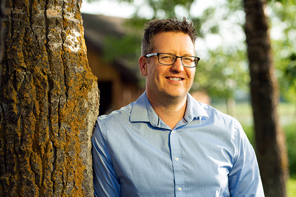 Photo of Clark Banack leaning against a tree and smiling at the camera.