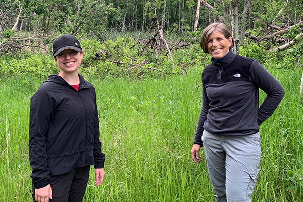 A photo of Glynnis Hood and Jacqueline Kublik standing in Miquelon Lake Provincial Park with grass and trees behind them.