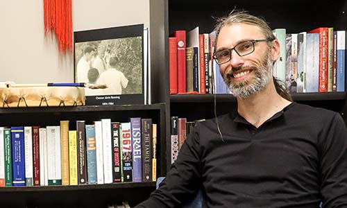 Photo of Joe Wiebe looking at the camera and smiling while sitting in front of black bookcases filled with books.