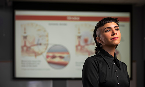 Photo of Ana Klahr looking off to the side with a slight smile on her face like she is pondering something. She is wearing a black button-up shirt and there is a slide of an illustrated model of the brain. 