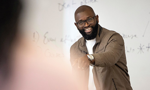 Photo of Michael Omoge in the front of a classroom smiling and gesturing to a blurred-out student in the foreground of the photo.