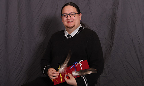 Photo of Gordon Naylor sitting in front of a black backdrop. His hair is pulled back and he is wearing glasses. He smiles at the camera as he is holding two eagle feathers and a small, rectangular board with four ribbons on it. The ribbons are each a different colour and represent the colours: blue, white, red, yellow.