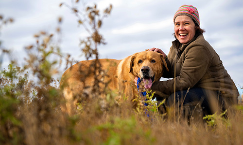 Photo Ivanna Schoepf in a field of golden grass, wearing a brown coat and knit toque while petting a gold-coloured dog.