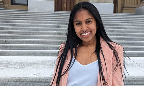 Jewel Anne Naicker standing on stone steps leading up to a tall, stone building. She has dark hair and is wearing a white shirt and pink blazer.