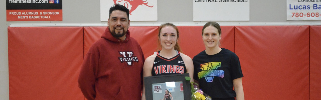 Lauren Cardinal in a Vikings basketball uniform in a gymnasium, holding a framed photo of herself playing basketball and a bouquet of flowers. She stands in between her father in a Vikings hoodie on the left and Vikings women's basketball head coach, Megan Wickstorm, who is in a Vikings t-shirt.
