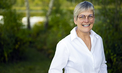 Dee Patriquin in a white button-down and wearing glasses as she smiles at the camera in front of a forested area.