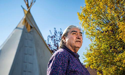 Elder John Crier standing outside and looking off into the distance, in the background is a tree and a tipi.