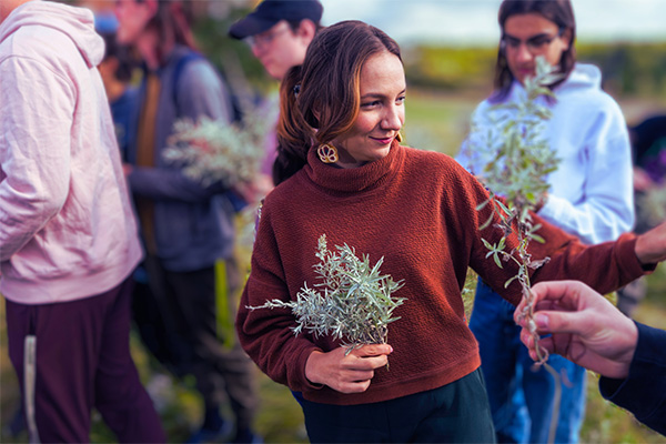 Faculty member Willow White standing in a grassy field with students while inspecting a sprig of sage.