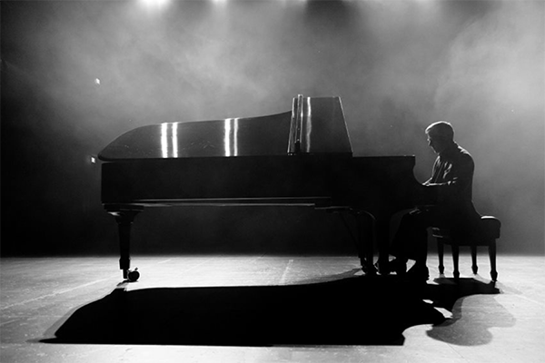 Black and white image of a man playing at a grand piano on a stage.