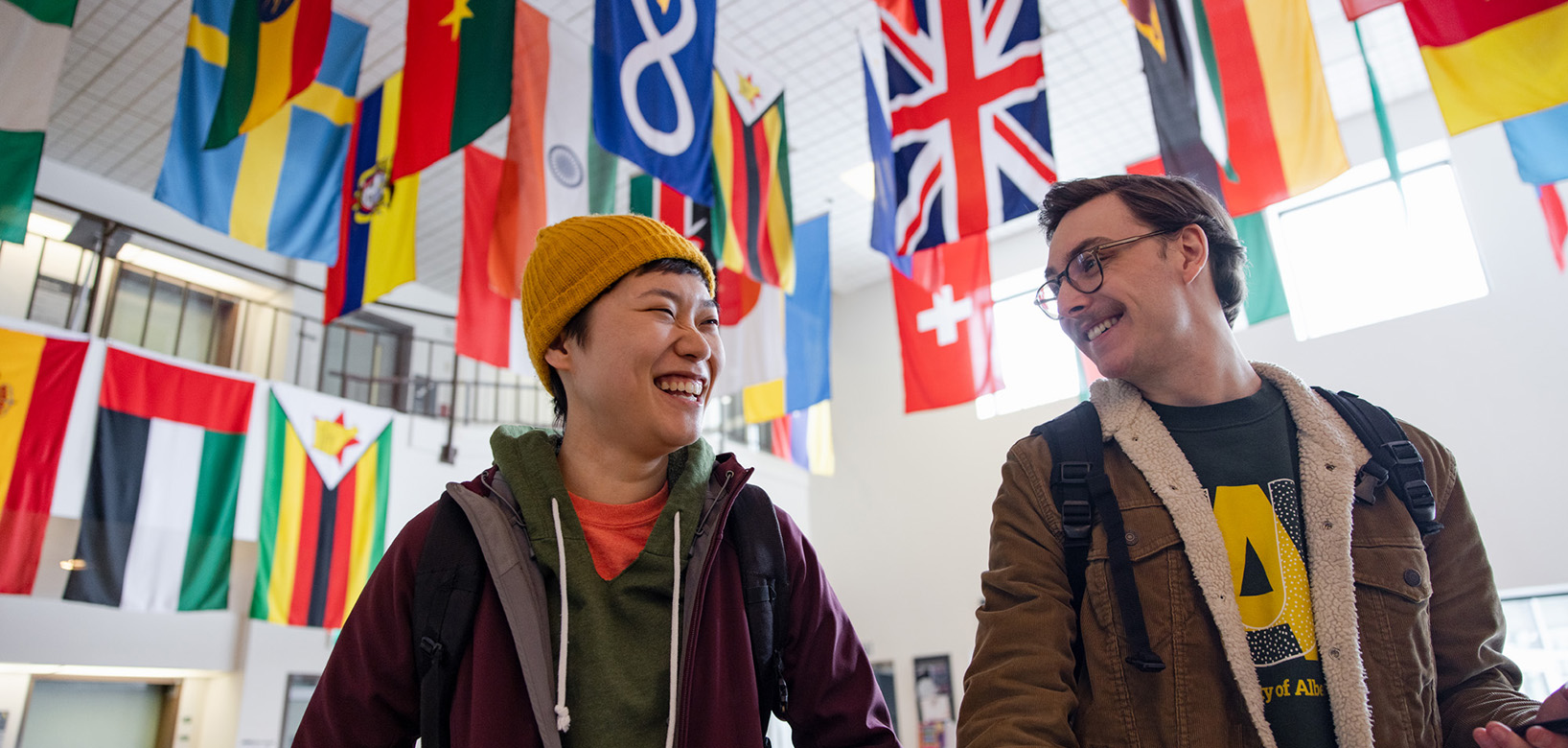 Two students standing in a room. Above them hand various flags from countries around the globe.