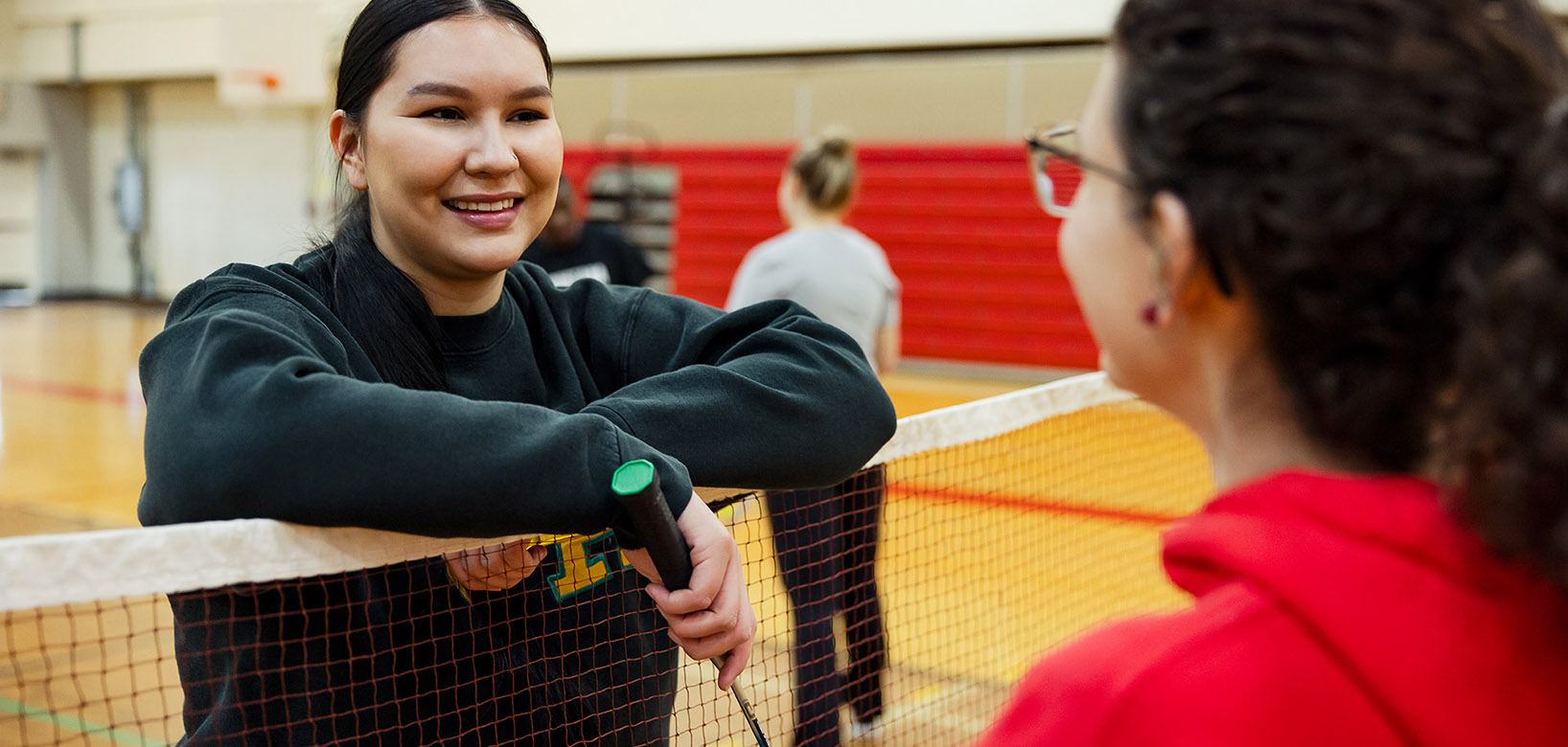 A student leaning on a badminton net as she talks with another student.