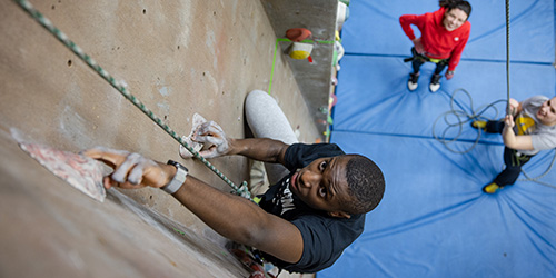 A student climbing a rock wall with two other students watching from below.
