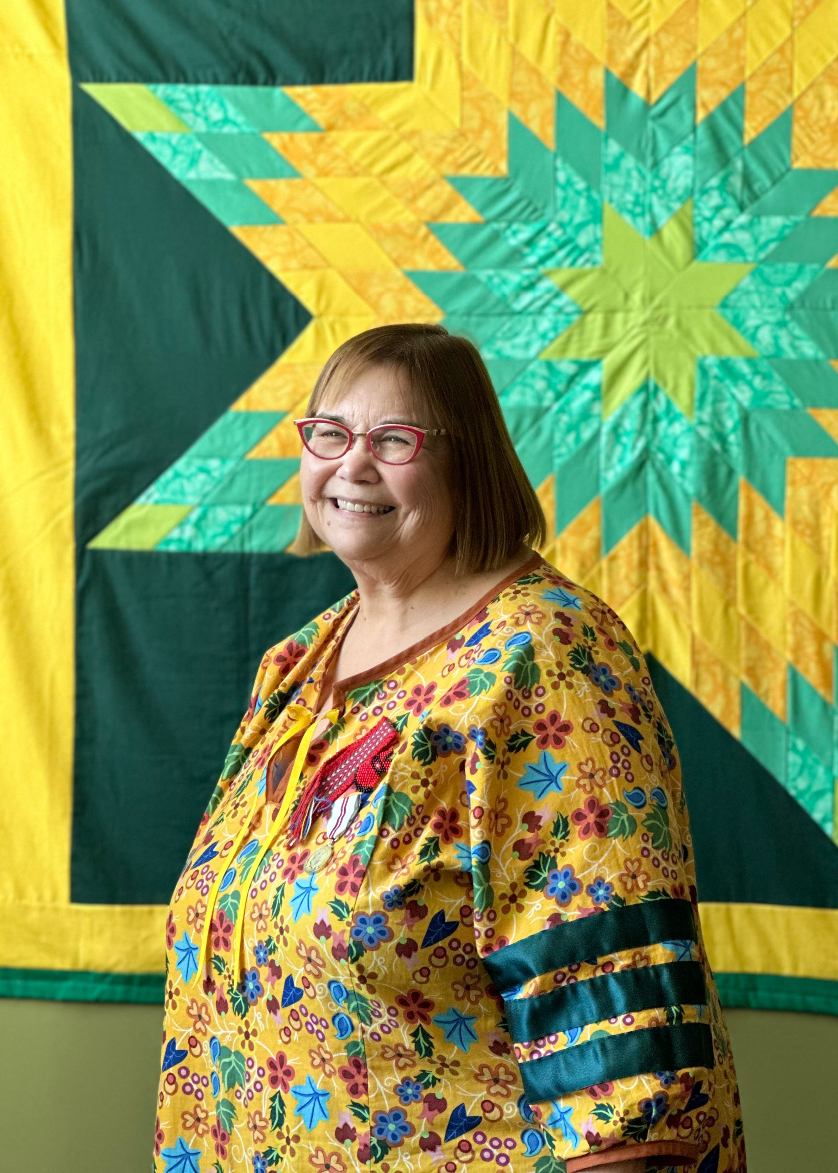 Dr. Florence Glanfield in yellow patterned dress standing in front of yellow and green quilt