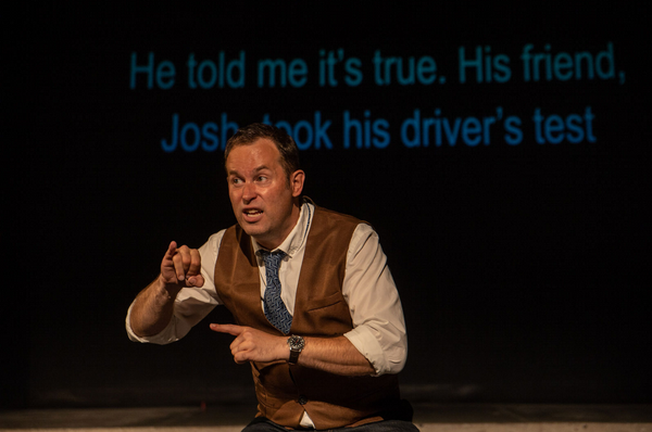 Actor Chris Dodd is onstage wearing a vest and tie, standing in front of a projection of a conversation that reads "He told me it's true. His friend Josh took his drivers' test."