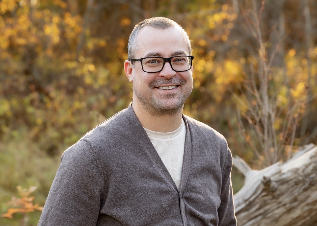 An image of Dr. David Rast, a man with glasses wearing a cardigan, standing near trees with yellow leaves