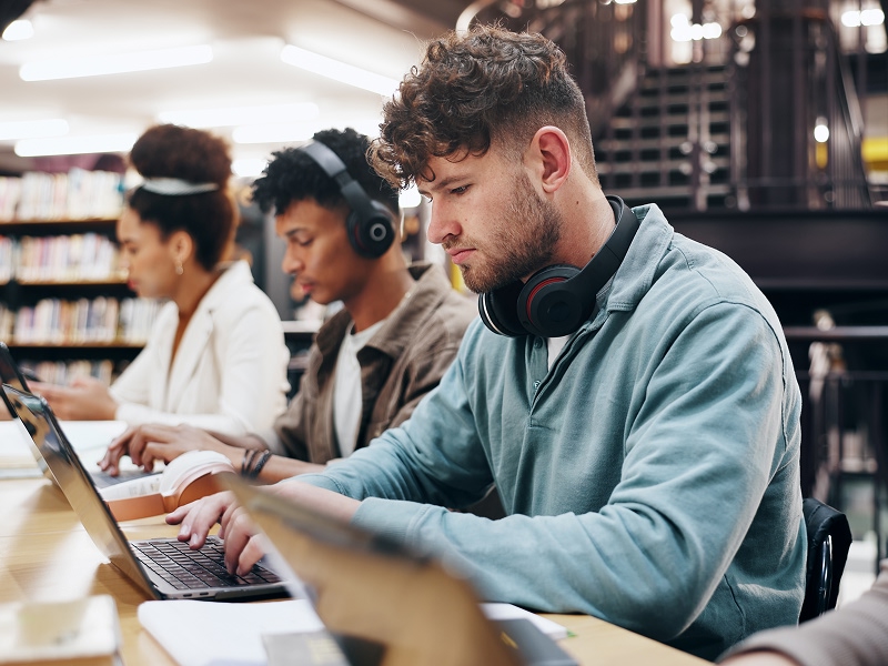 Students studying on laptops in a library, with headphones and books on the table.