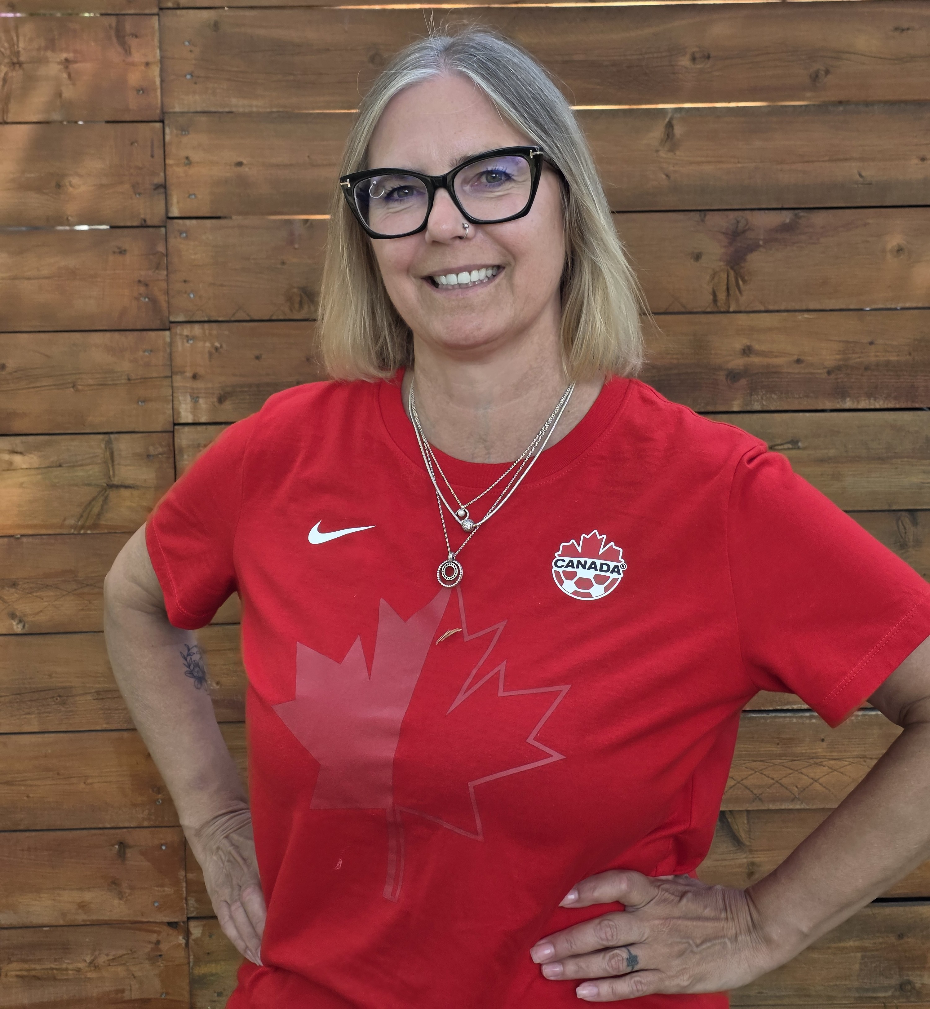 An image of Dr. Wendy Hoglund, a woman wearing a Team Canada soccer jersey while standing in front of a wooden fence.
