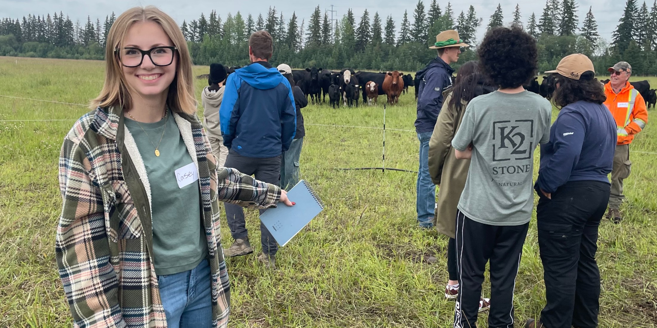 A students standing in a field with a clipboard, with livestock and people in the background.