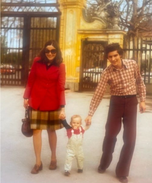 A smiling couple in retro attire — the woman in a red coat and plaid skirt, the man in a patterned shirt and flared pants — hold a toddler’s hands as the child takes steps in front of a decorative iron gate.