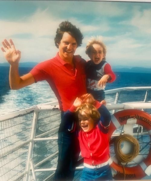 A man in a red shirt smiles and waves while holding two young children on a ferry deck, with ocean water and distant shoreline visible behind them.
