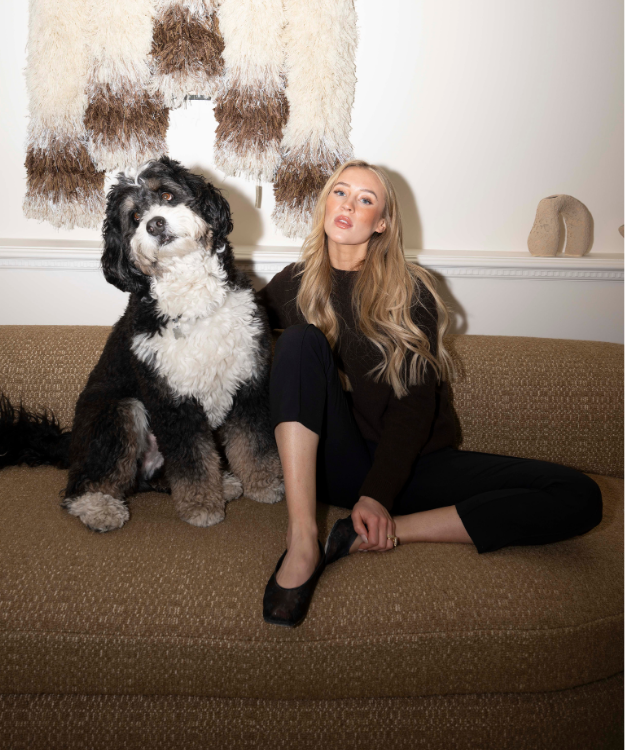 Lauren Kyle McDavid sits on a neutral-toned sofa beside her large black-and-white dog named Lenard, with a textured wall hanging and minimal decor in the background.