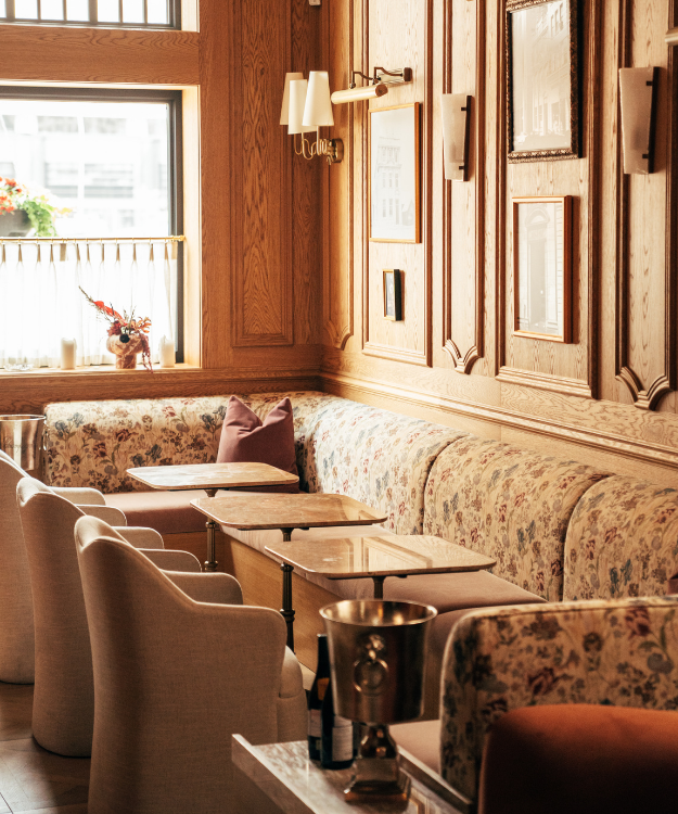 Bar Trove interior: A softly lit lounge with wood-paneled walls, framed artwork and sconces, featuring floral banquette seating, marble tables and upholstered chairs arranged along a window.