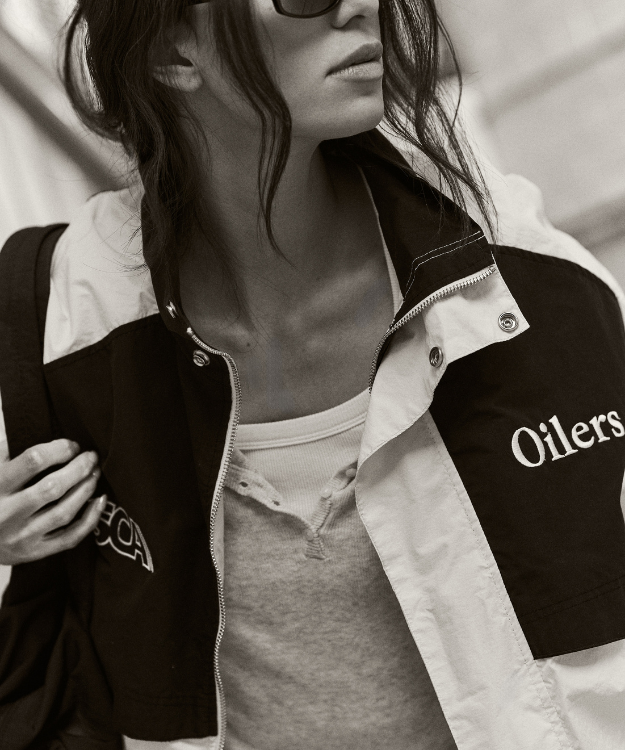 Close-up of a model in a Sport Club Atelier Oilers jacket, styled with layered basics and sunglasses in a monochrome shot.