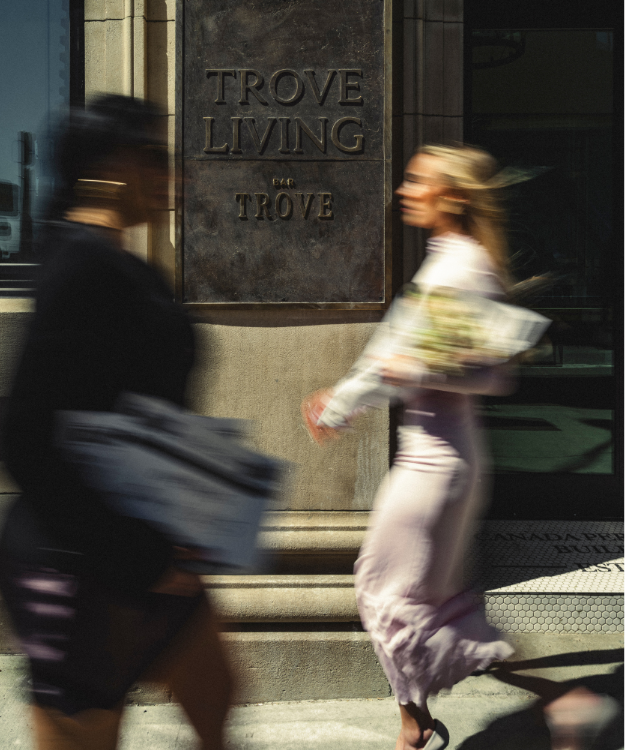 Blurred figures walk past the Trove Living and Bar Trove sign on a city building, capturing a sense of motion.