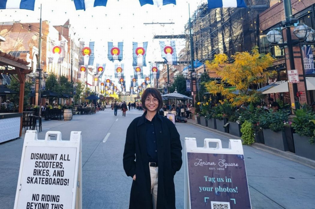 A person smiling on a pedestrian street in Larimer Square, Denver, with Colorado flags strung above outdoor patios.