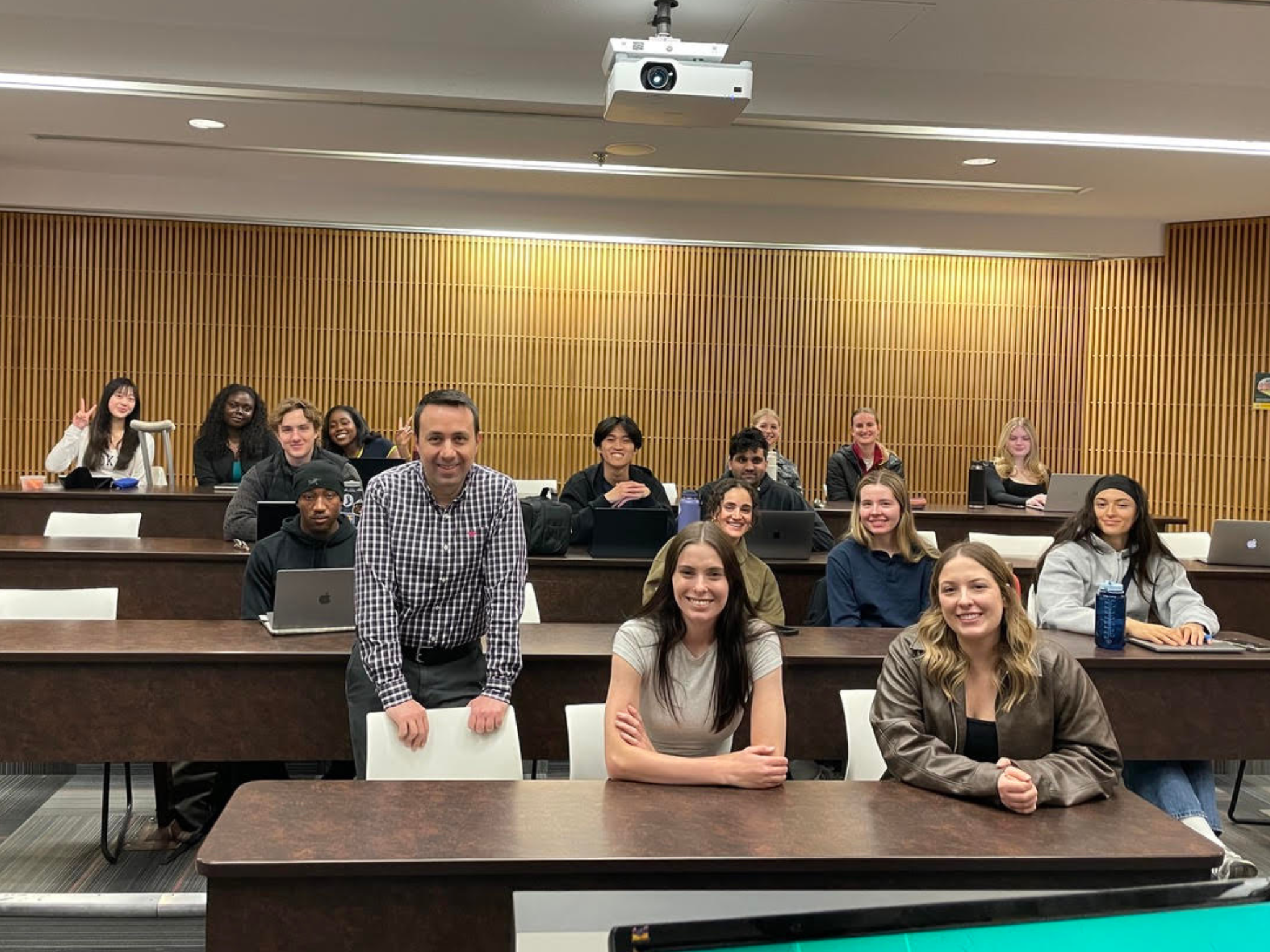 Students in a University of Alberta classroom sit in tiered rows with laptops open, as an instructor stands at the front with two students, all facing the camera.