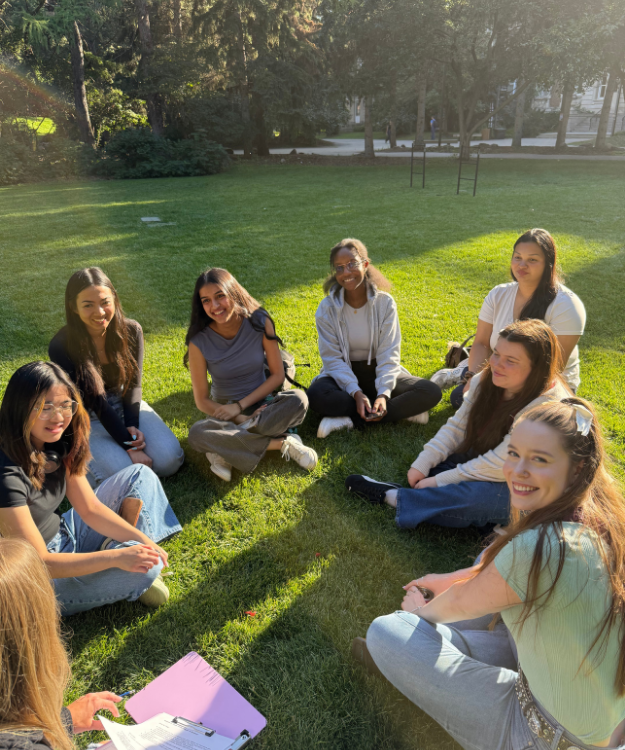Members of the Women in Business student group at the University of Alberta sit in a circle on a grassy lawn, talking and smiling in the sun.