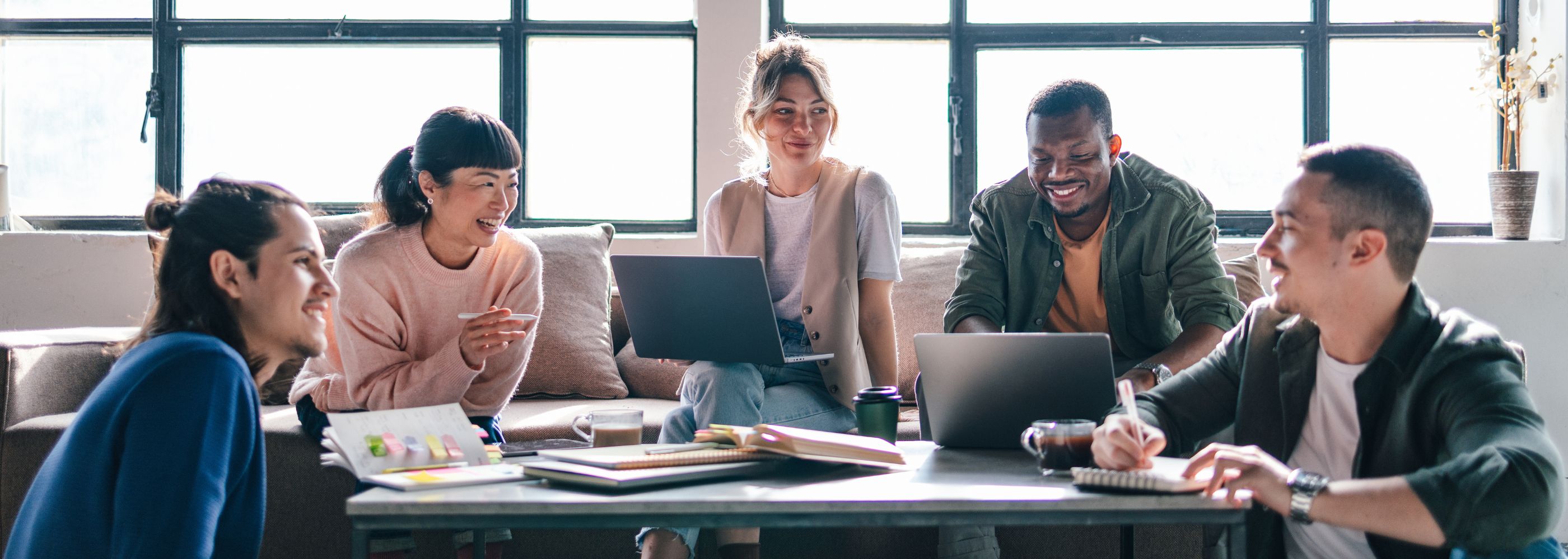 A multicultural group of young adults engage in teamwork at a modern co-working space with laptops and smiles.