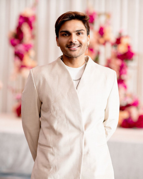 Headshot of Ibrahim Syed: A person with short dark hair smiles at the camera, wearing a cream-coloured tailored jacket, layered necklaces and a drop earring, standing in front of a softly blurred backdrop of pink and peach floral arrangements.