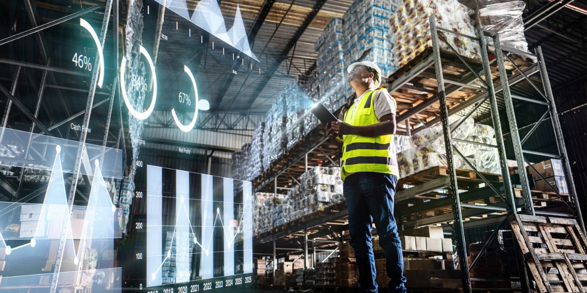 Worker in a high-visibility vest using a tablet in a warehouse aisle, with overlaid charts and graphs representing real-time supply chain and inventory data.