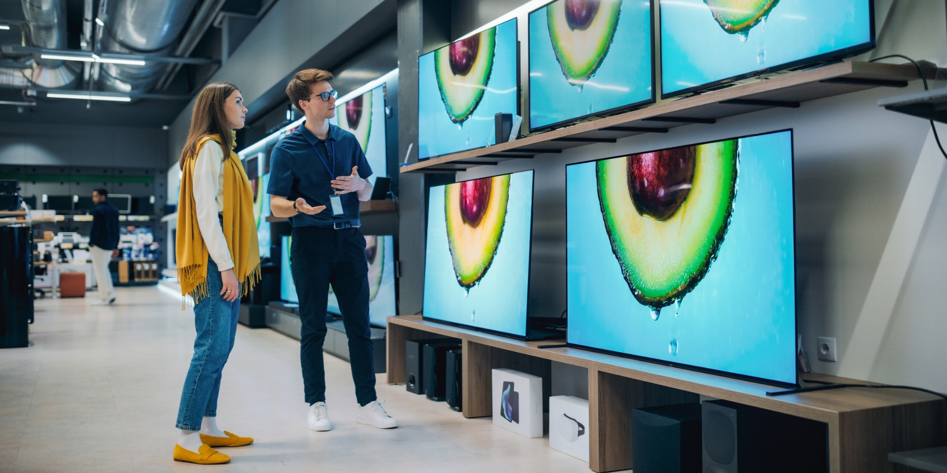 A customer and sales associate compare televisions in an electronics store, surrounded by large screens displaying vibrant images.