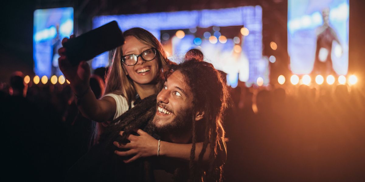 Two people at a nighttime outdoor concert take a selfie in front of a brightly lit stage, smiling as a crowd surrounds them.