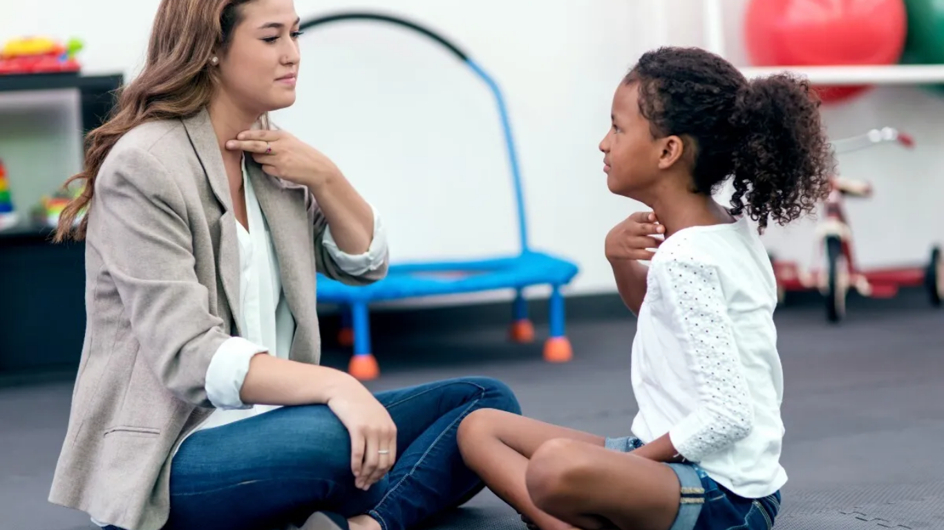 Therapist sits with child holding fingers to their necks during a speech assessment