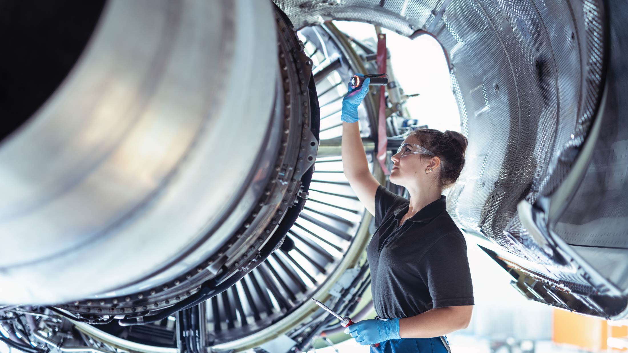 A female apprentice aircraft maintenance engineer works underneath jet engine.